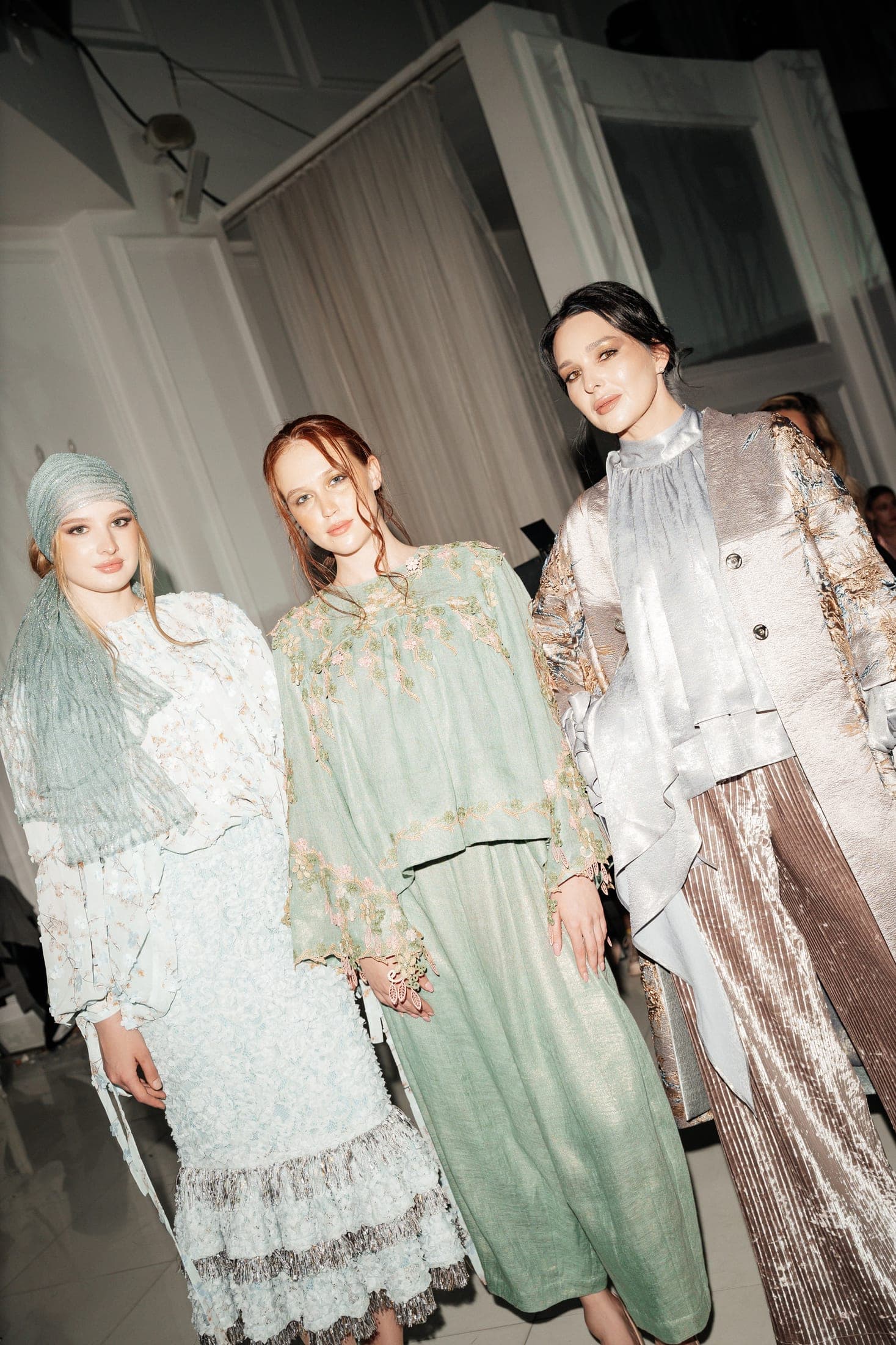 Three models backstage in pastel embellished looks — a mint embroidered set, a pale blue floral blouse and a metallic brocade coat
