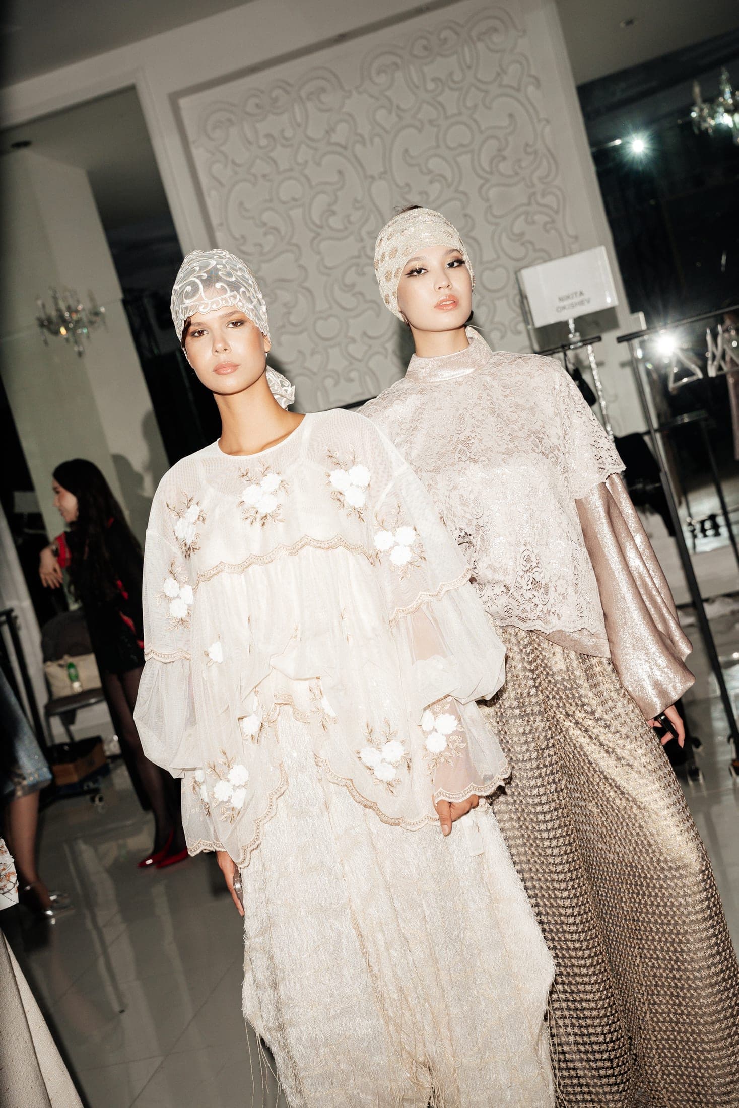 Two models backstage in ivory lace headpieces, wearing a floral-appliqué blouse with a fringed skirt and a lace top with a gold houndstooth skirt