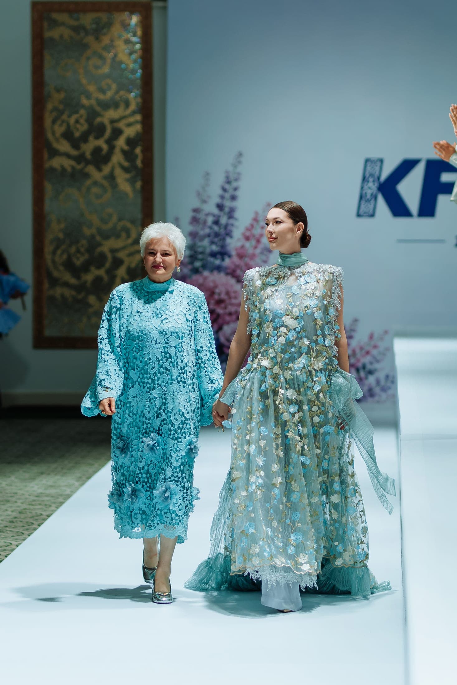 The designer and a model in turquoise gowns walking hand in hand at the runway finale