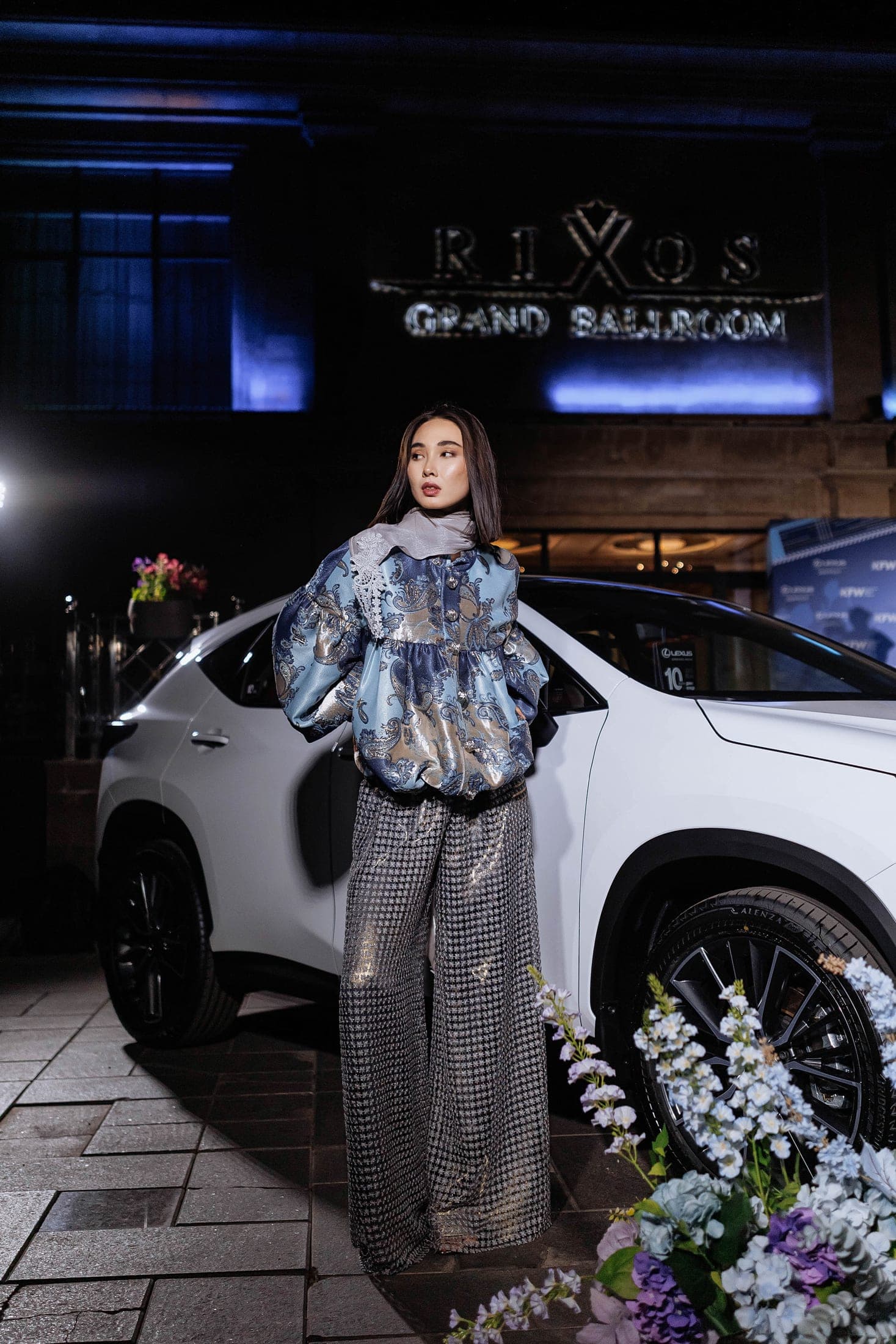 Model in a blue paisley brocade jacket and sequined trousers posing beside a white Lexus outside the Rixos Grand Ballroom