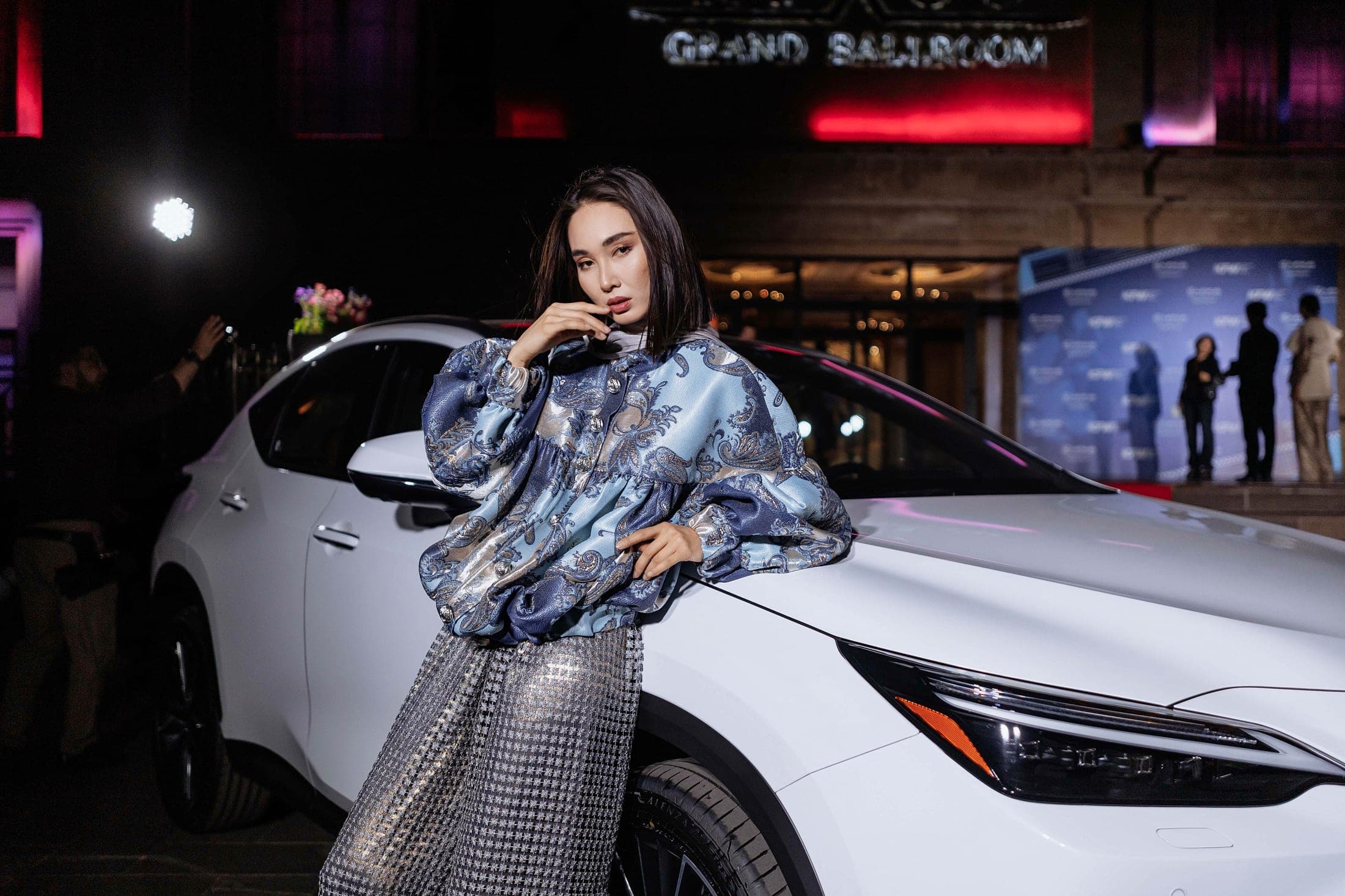 Model in a blue paisley brocade jacket leaning against a white car at night