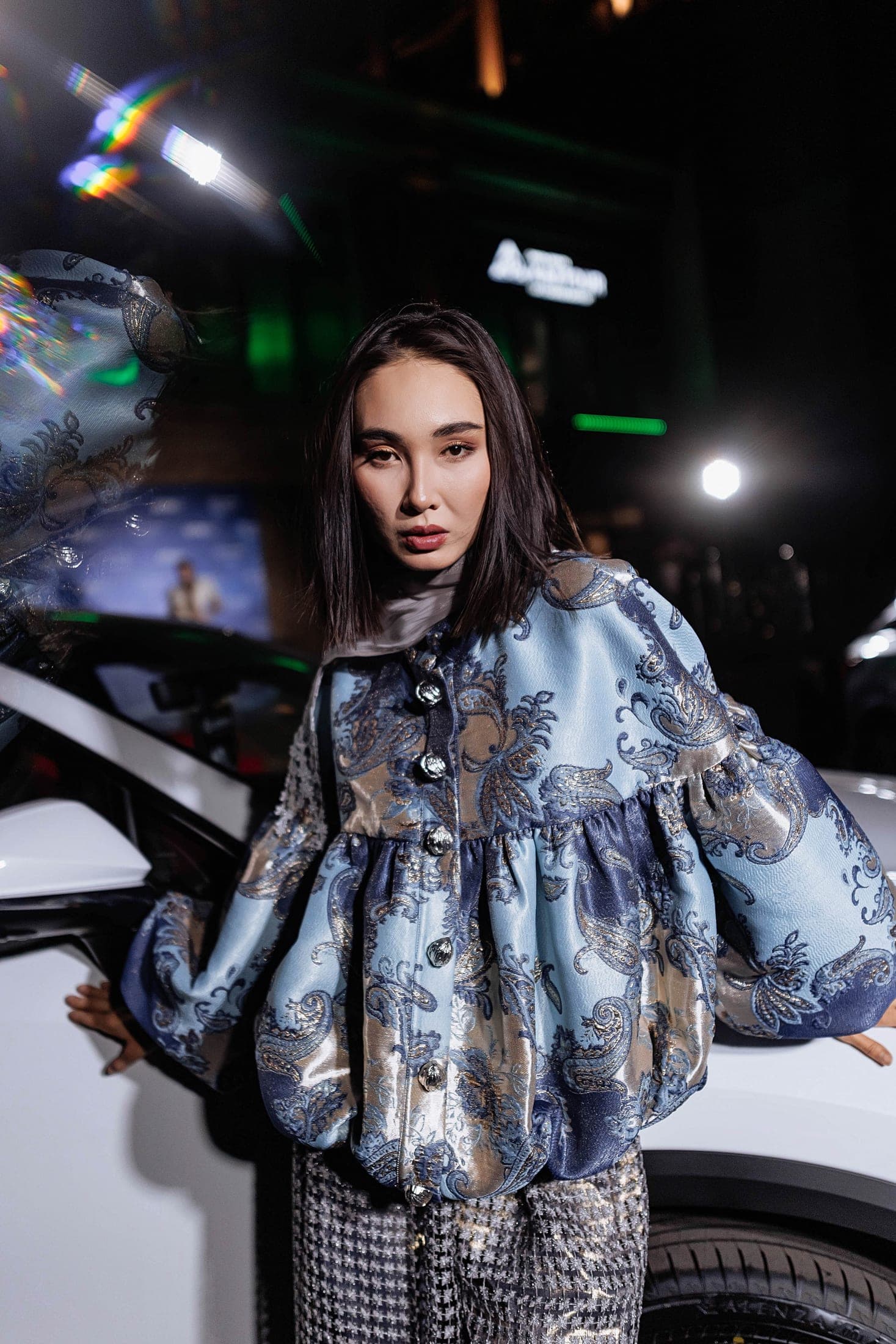 Portrait of a model in a blue paisley brocade jacket with a pale scarf beside a white car at night