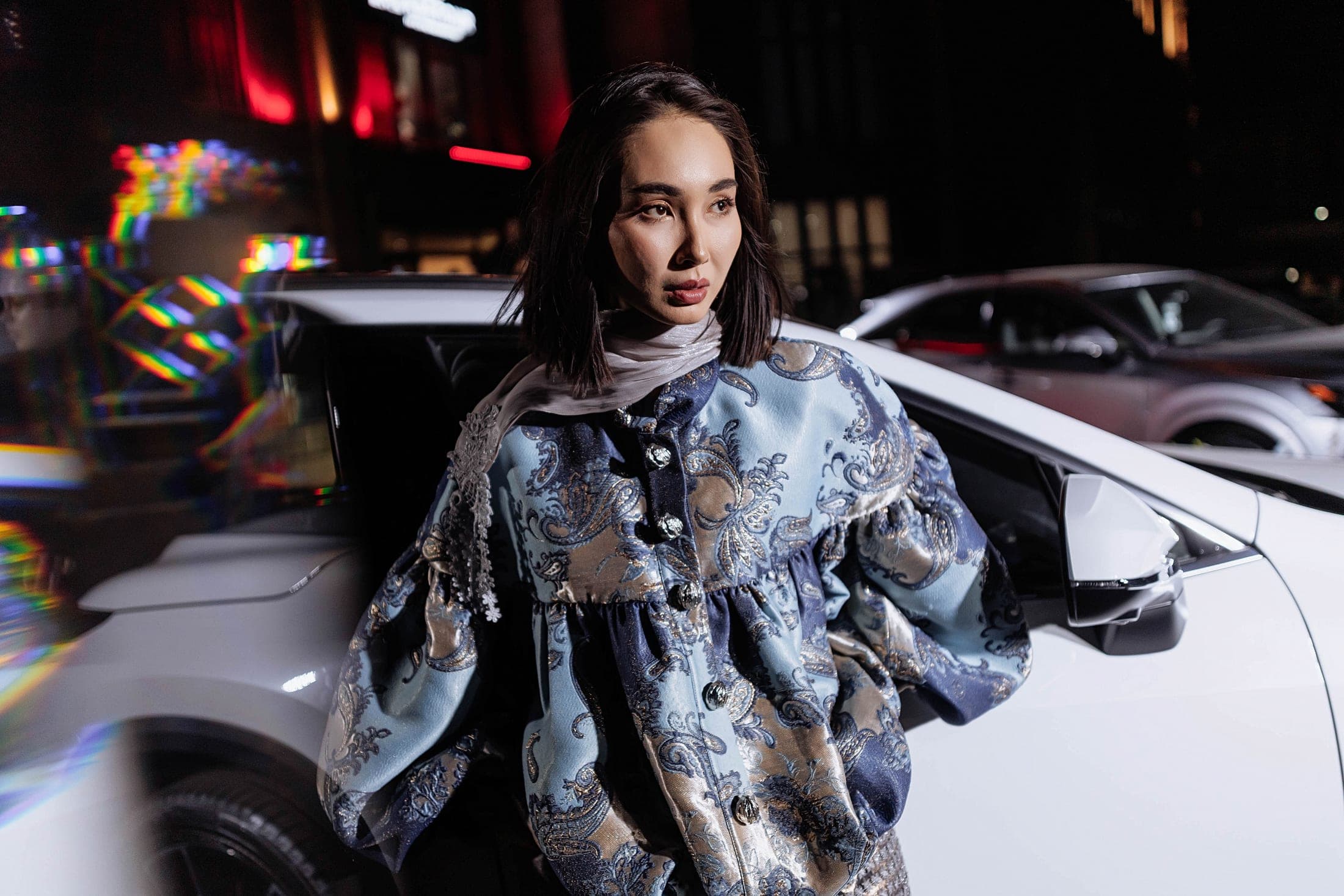 Model in a blue paisley brocade jacket beside a white car, prism light flares across the frame