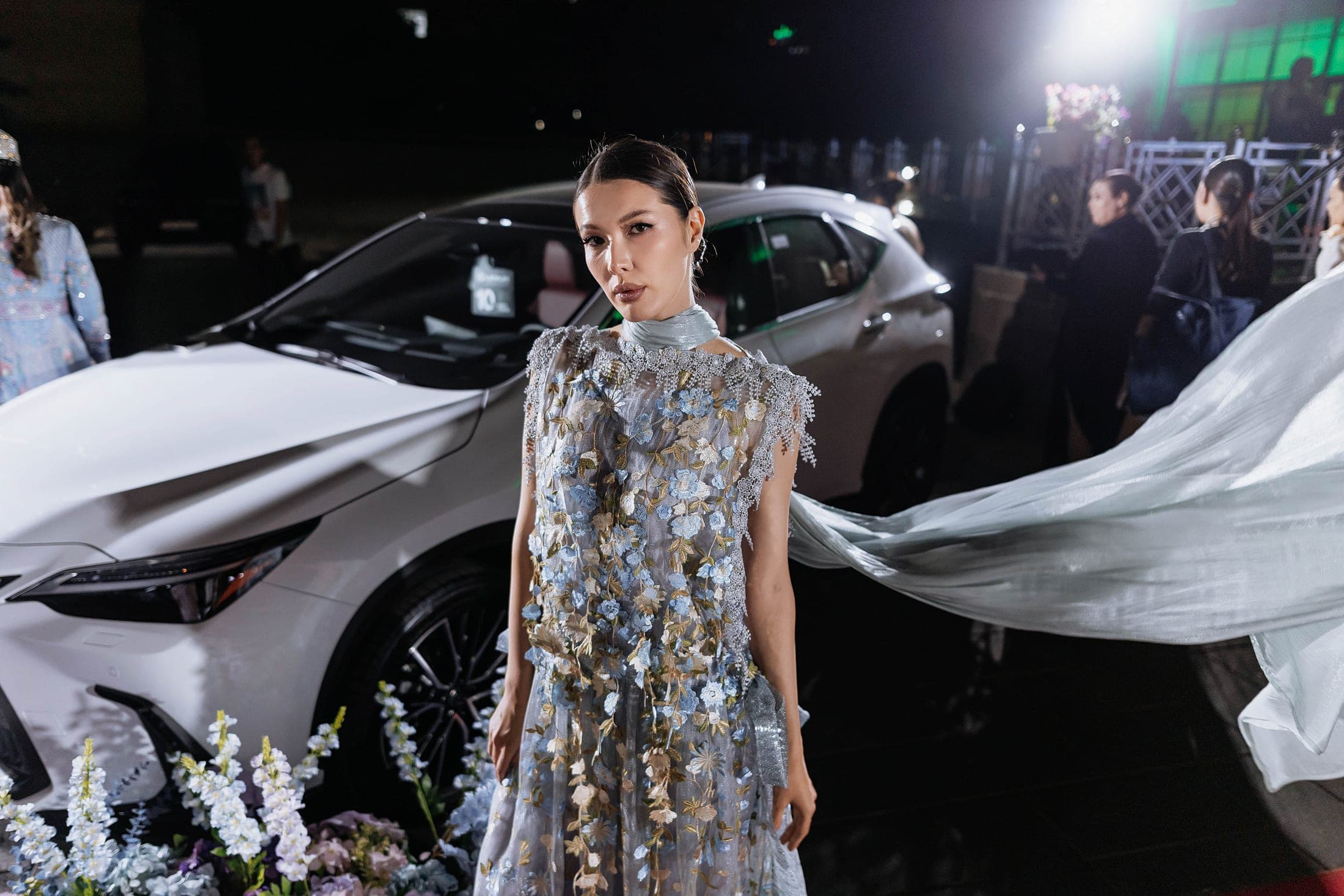 Model in a pale blue floral-embroidered dress with a flowing white veil beside a white Lexus