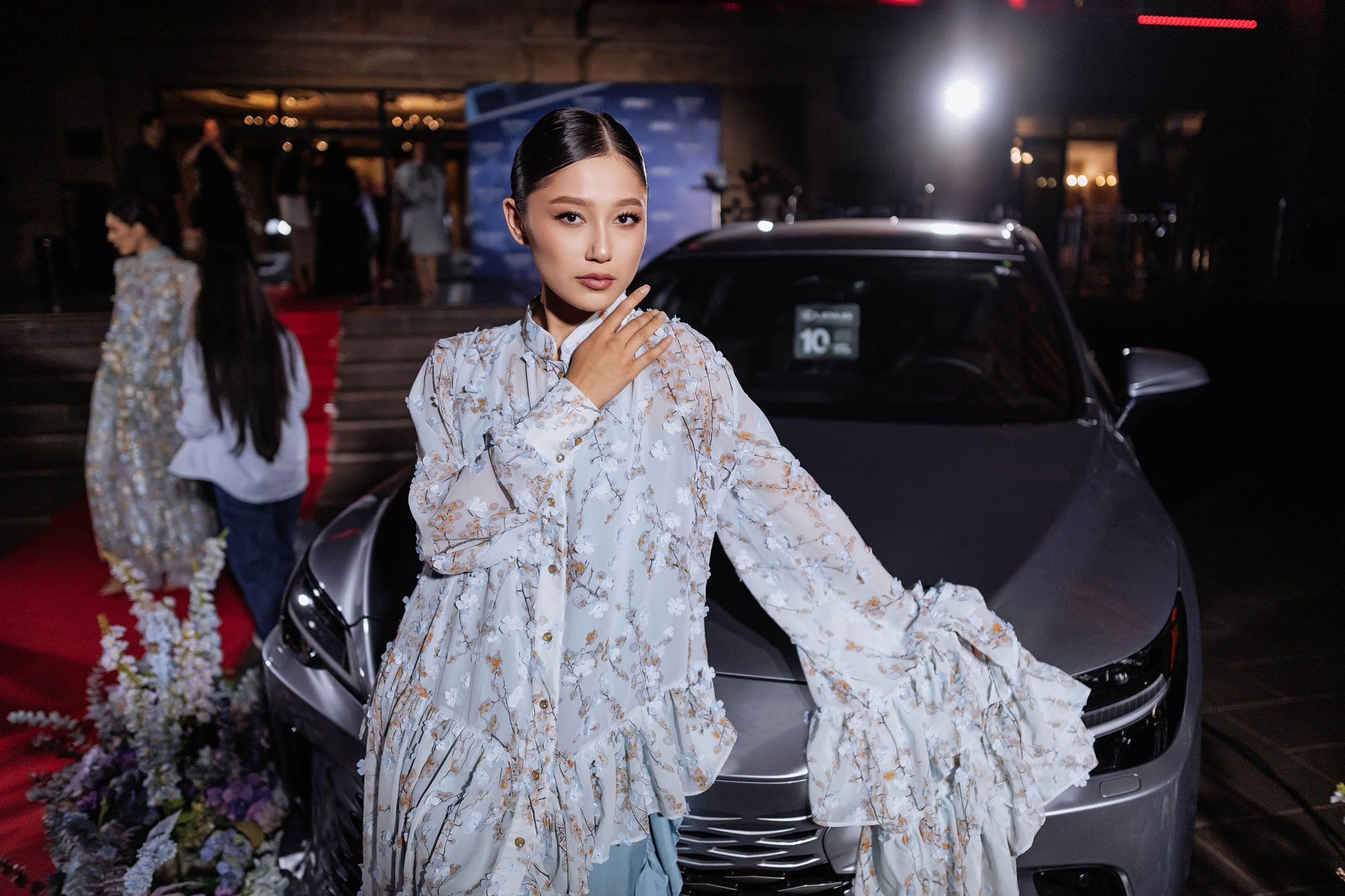 Model in a blossom-appliqué chiffon blouse beside a grey Lexus at the red-carpet arrivals
