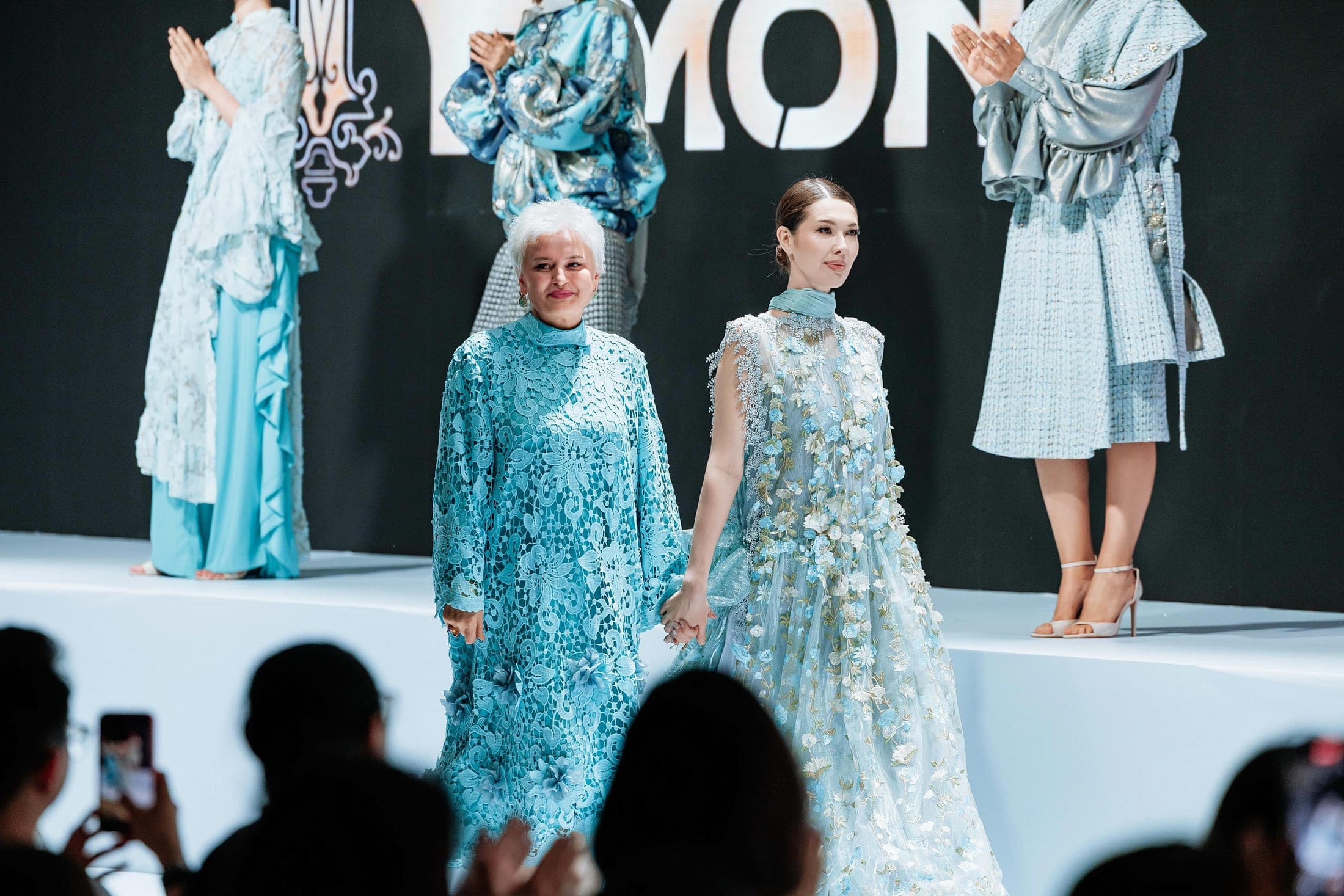 Two models on the runway — a blossom-print chiffon dress and a paisley brocade jacket — before the KFW Shymkent backdrop