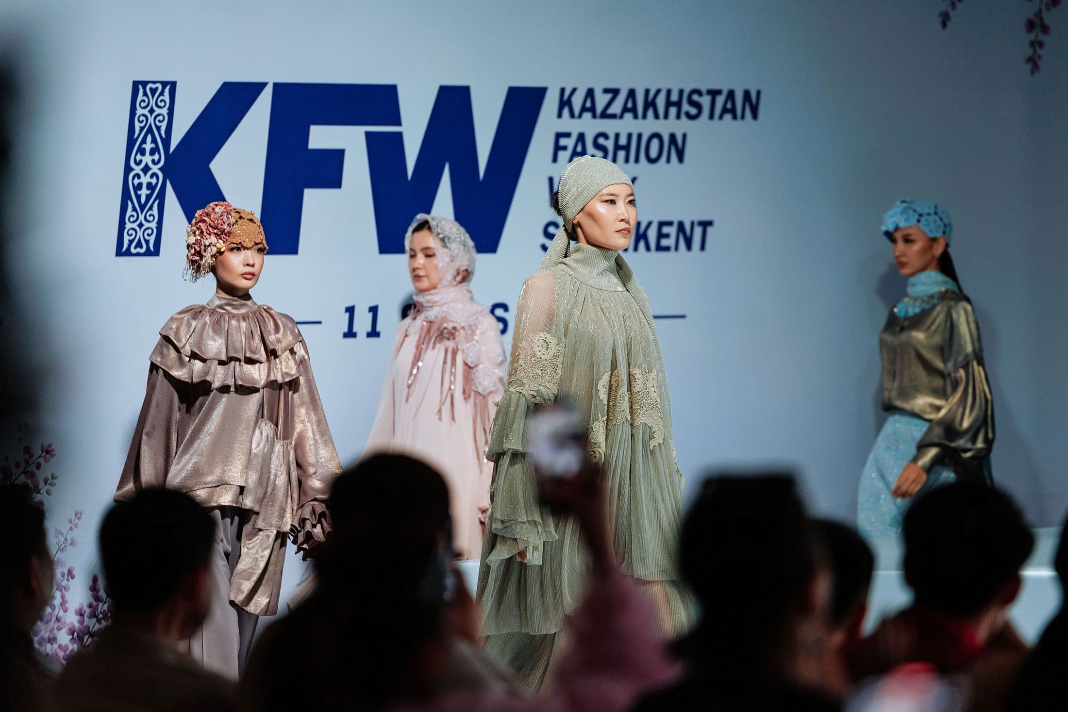Models on the runway in ivory feather-collar, gold metallic and sage-green looks before the KFW backdrop