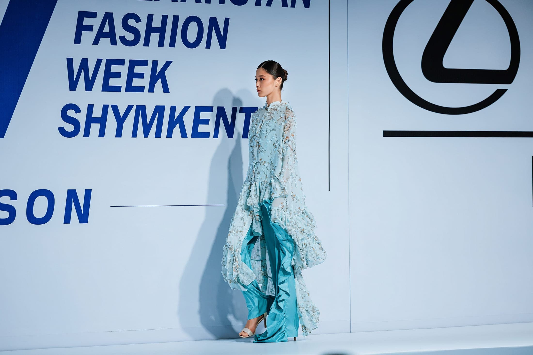 Model walking the runway in profile in a pale blue blossom-appliqué dress over turquoise silk trousers