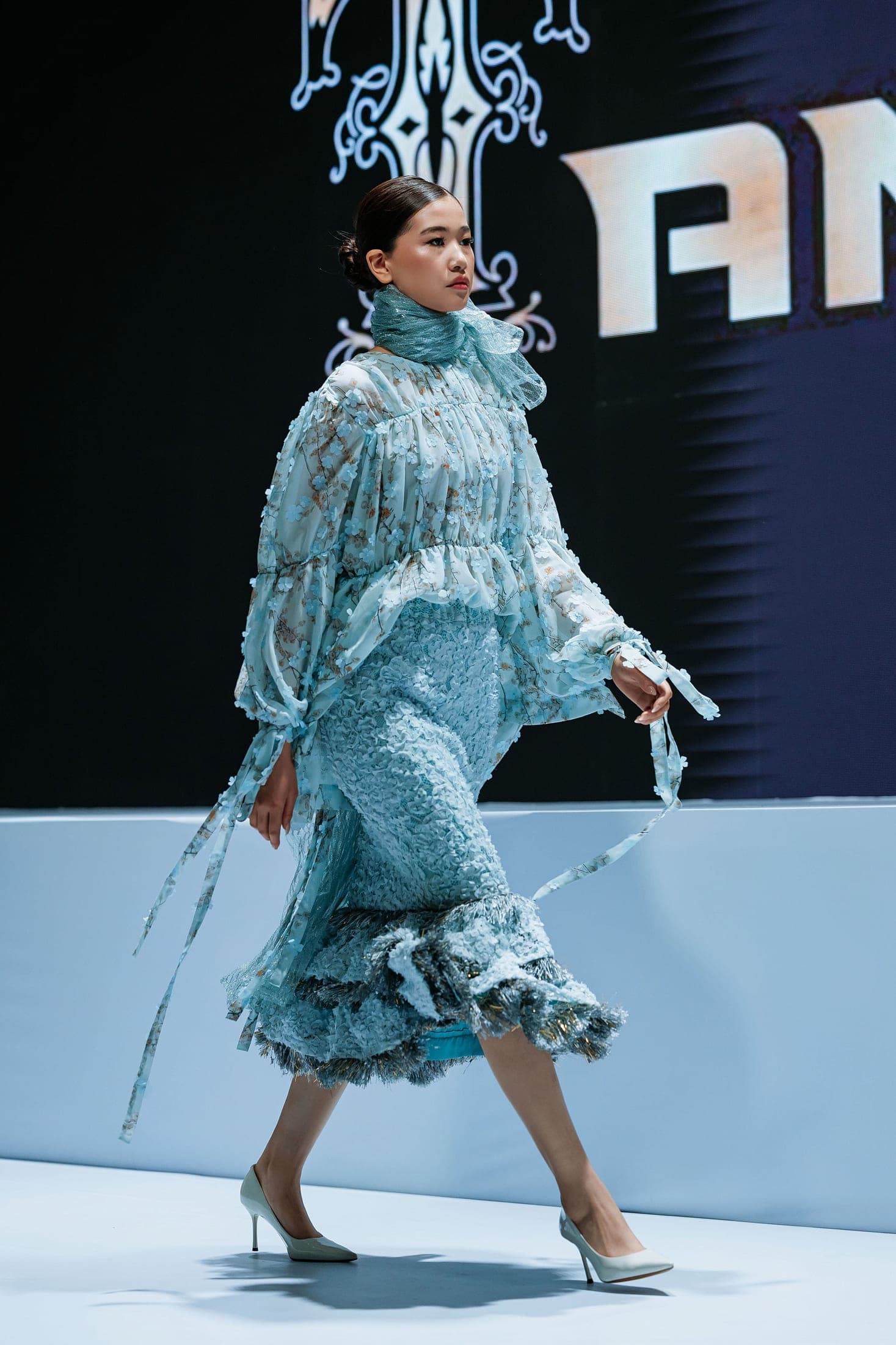 Model on the runway in a blossom-print ruffled blouse and textured lace skirt before the Tamone backdrop