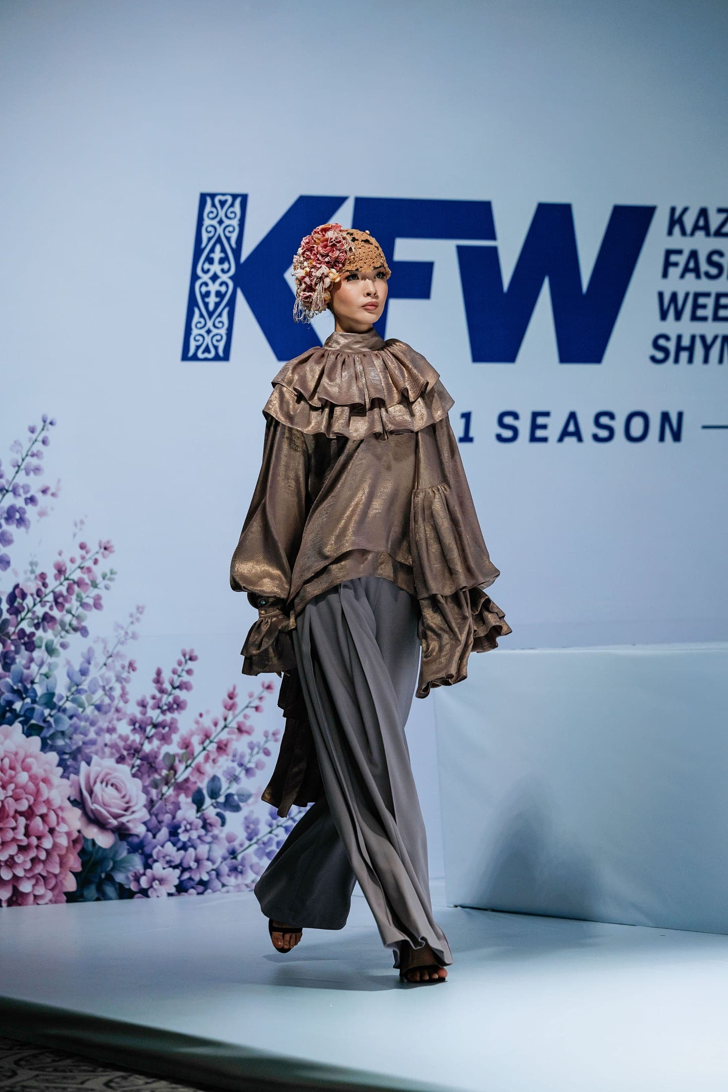 Model on the runway in a bronze ruffled blouse and draped grey trousers with a floral crochet headpiece