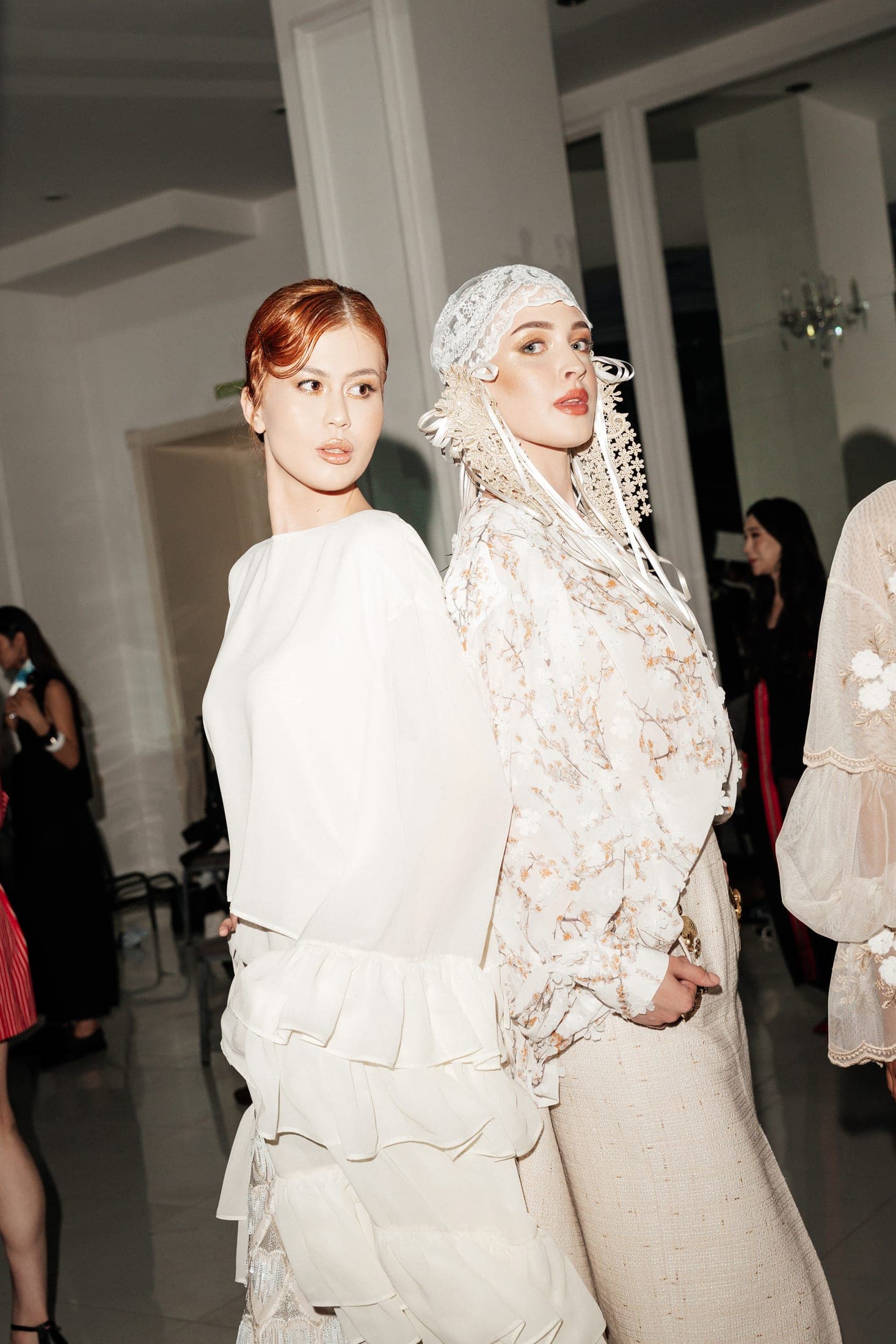 Two models backstage, one in an ivory ruffled blouse, the other in a blossom-print blouse with a lace bonnet