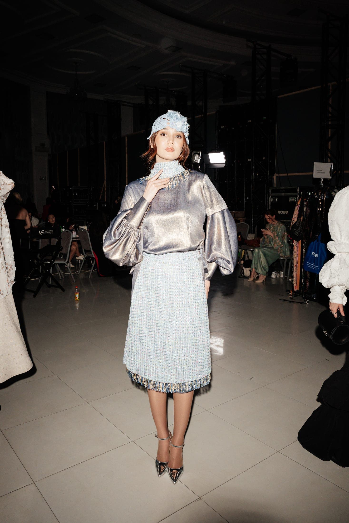 Model backstage in a metallic silver blouse, floral headpiece and pleated skirt