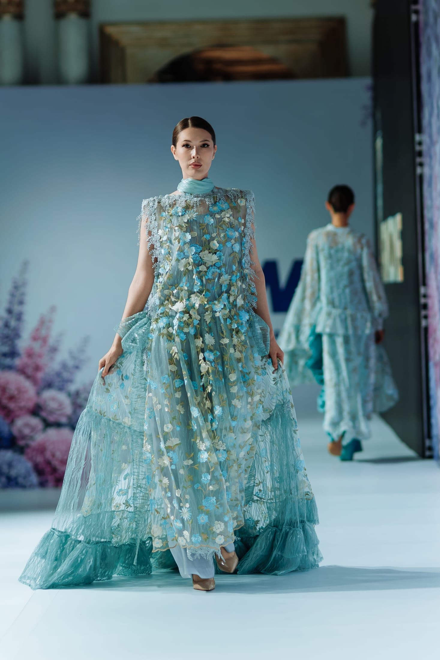 Model walking the runway in a sheer turquoise floral-embroidered gown with pearl trim, another model following behind