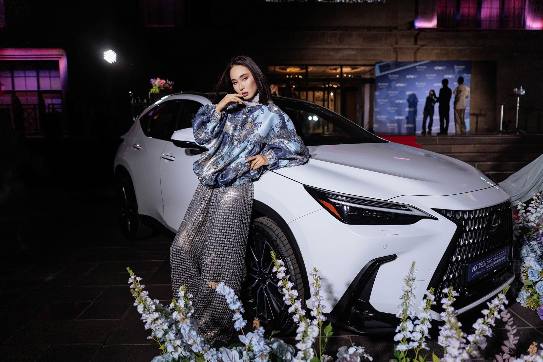 Model in a blue paisley brocade jacket and sequined trousers posing beside a white Lexus