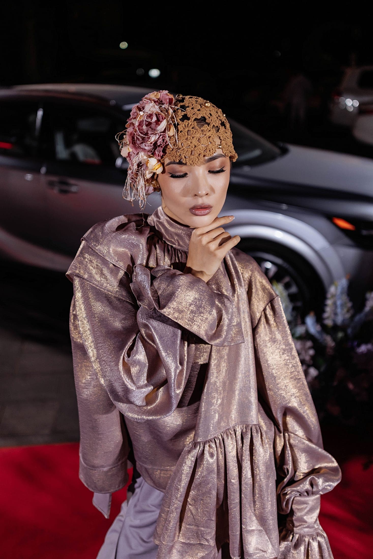 Model in a metallic rose-gold ruffled dress and dried-flower headpiece beside a car at night